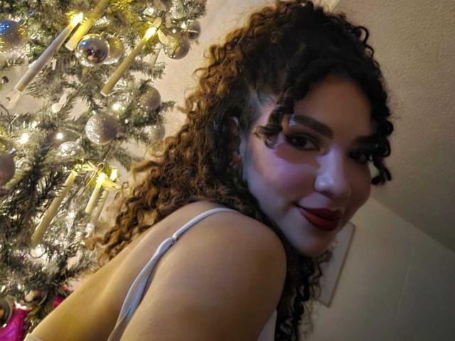 KendallPiierce69 with curly brown hair, white tank top, red lipstick, smiling at camera, Christmas tree with candles and ornaments in background, warm indoor lighting.