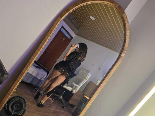 KendallPiierce69 with curly hair, wearing a black lace top and short skirt, showing her ass in a mirror selfie, holding a phone, in a room with wooden ceiling.
