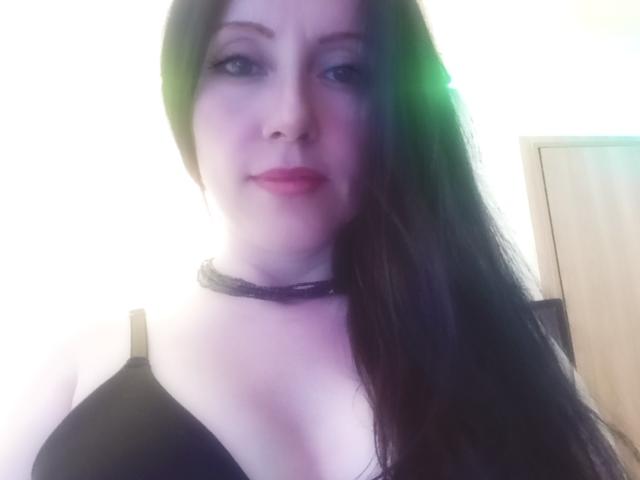 MondBlue has pale skin, long black hair, wearing a black bra and a black choker. Boobs are visible, she's smiling with pink lipstick. Light green lens flare on left.