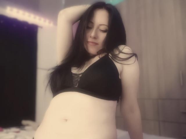 MondBlue with long black hair, wearing a black bra, has her arm raised, showing her pale skin and small tits, in a blurry bedroom with soft lighting.