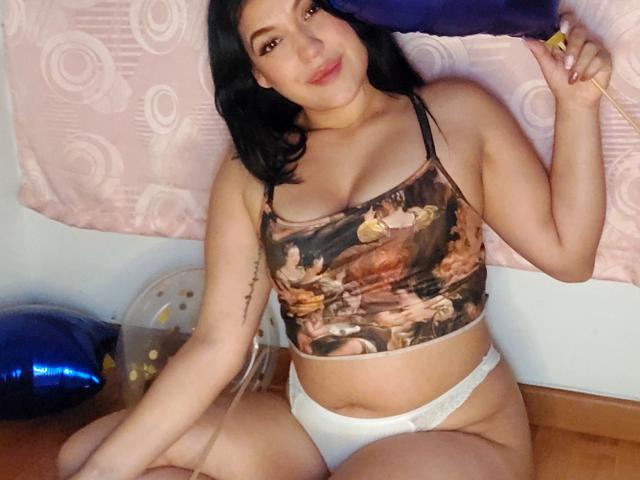 LauraRichis with black hair, wearing a camouflage sports bra and white panties, sitting on a wooden floor, holding a yellow object, smiling, showing her tits and ass.