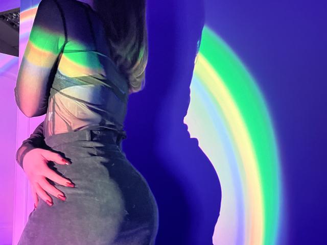 IsleBelle's ass in a tight grey dress, hands on hip, rainbow light on blue wall, and red nails. Her back is to the camera, showing her head silhouette.
