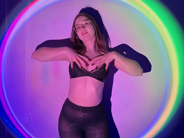 IsleBelle con gafas, un sujetador de encaje negro y pantalones negros transparentes, con las manos en el pecho, de pie, con una luz de anillo de colores, tonos púrpura y verde, segura de sí misma, sexy, delgada, cabello castaño..