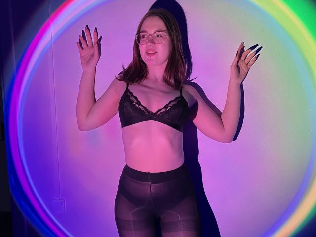 IsleBelle with glasses, black lace bra, and sheer black pants, standing with hands up, colorful ring light behind, smiling, fair skin, brown hair, small tits, visible nipples.