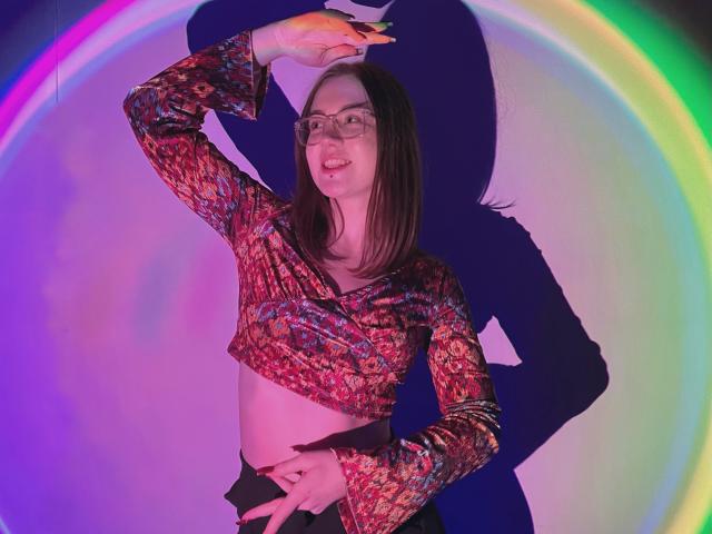 IsleBelle with glasses, medium tits, brown hair, red patterned crop top, black pants, arm raised, shadow on wall, colorful ring light, smiling, right hand on belly.