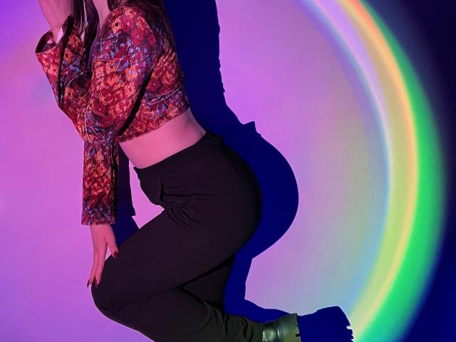 IsleBelle with colorful patterned top, black pants, and green boots, kneeling with a rainbow light effect on the right, showing her ass and a shadow on the blue wall.