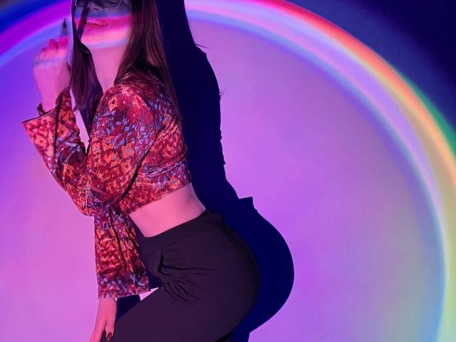 IsleBelle with long brown hair, wearing a colorful patterned top and black pants, holding a microphone, illuminated by a rainbow light effect, her ass slightly raised.