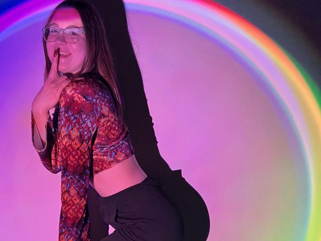 IsleBelle with glasses, smiling, hand to mouth, wearing a colorful patterned top and black pants, standing in front of a rainbow light, showing her ass.