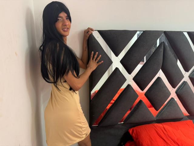 LatinMia, avec de longs cheveux noirs, souriante, portant une robe jaune, touchant une tête de lit grise avec un motif en forme de X, debout à côté d'une draps rouge..