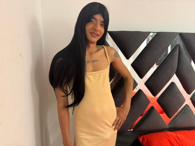 LatinMia: Capelli lunghi e neri, vestito giallo, seno piccolo, petto tatuato, mano sul fianco, sorridente, in piedi di fronte a una testiera scura con motivi, cuscino rosso..
