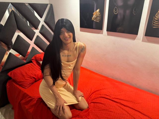 LatinMia, cheveux longs et noirs, robe jaune, tatouage sur la poitrine, agenouillée sur un lit rouge, souriante, mains sur la robe, chambre avec une tête de lit noire et des œuvres d'art au mur..