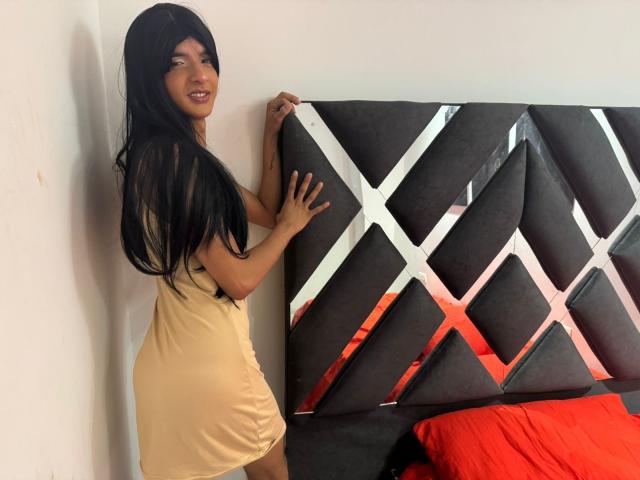 LatinMiaHot with long black hair, wearing a short beige dress, standing against a dark gray, diamond-patterned headboard, smiling, showing her ass, and holding the headboard with one hand.