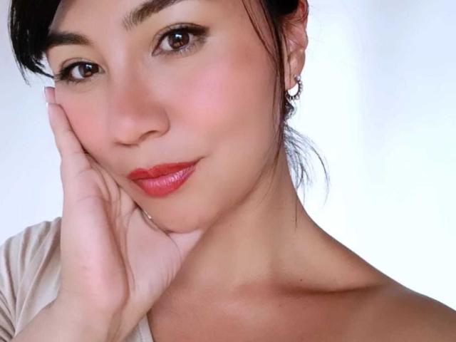 CharlotteBliss has her hand on her cheek, brown eyes, red lipstick, and dark hair in a ponytail, with a soft smile and subtle makeup, showing her bare shoulders.