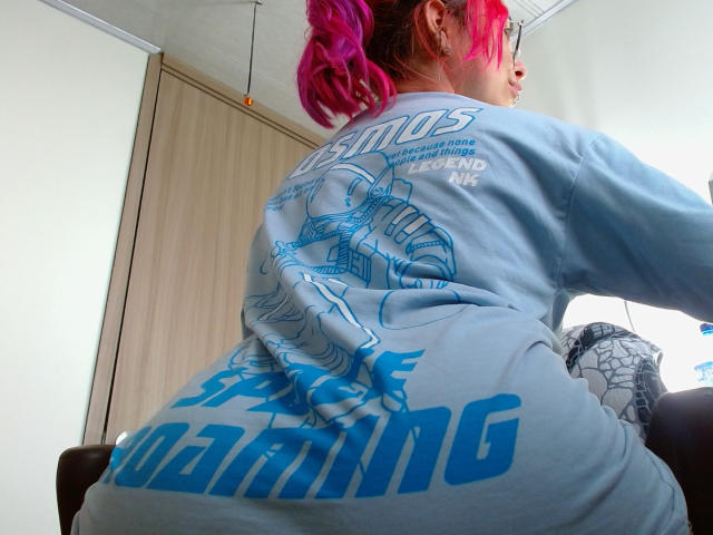 NatashaLau with bright pink hair, back to camera, wearing a blue graphic hoodie, sitting, showing her ass, in a room with a wooden door.