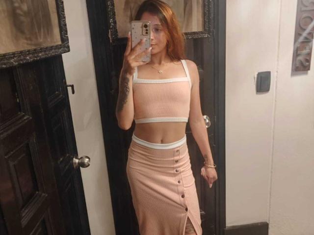 SaarahJones with red hair, wearing a pink ribbed crop top and matching high-waisted skirt with buttons, showing a tattoo on her right arm, holding a phone.