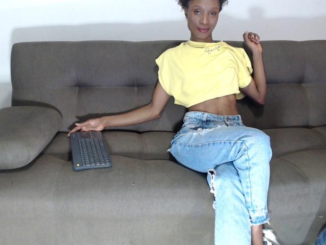 DesireHunterr with small tits, wearing a yellow crop top and ripped jeans, sitting on a gray couch with a keyboard, holding her shirt with one hand.