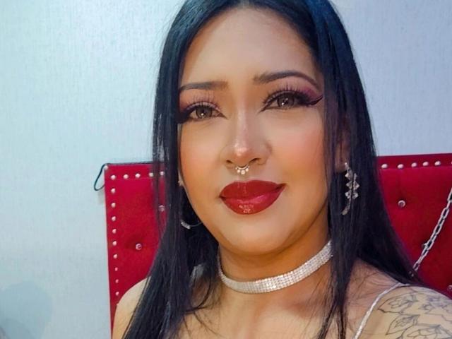 MelanieMorel with long black hair, red lipstick, nose ring, and chandelier earrings, wearing a sparkly necklace, smiling at the camera, red headboard in the background.