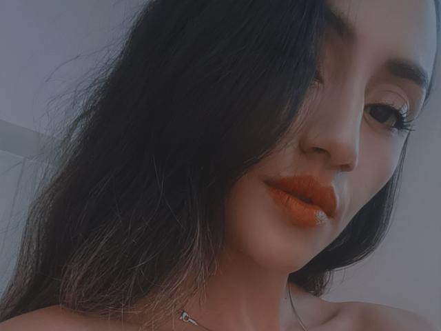 Helen Madisson, avec ses cheveux foncés, légèrement en désordre, son rouge à lèvres orange et son petit nez, regarde sur le côté avec un sourire discret. Ses cils sont longs et elle porte un collier..
