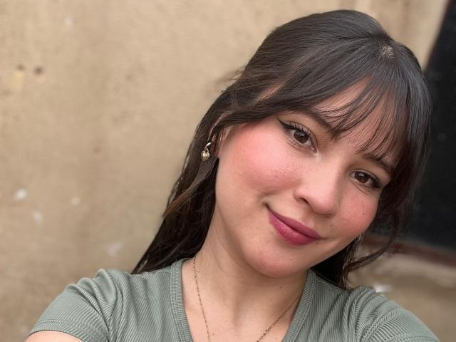 CelesteDash has long brown hair, bangs, and a cute smile. She's wearing a green ribbed shirt, a gold necklace, and small earrings. Her pinkish skin is slightly flushed.