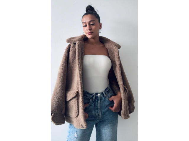 ThaliaAres with dark skin, in a brown fuzzy coat, white strapless top, and blue jeans, hands in pockets, hair in a bun, hoop earrings, looking down.
