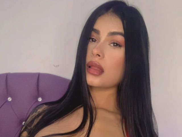 Cylielee with long black hair, pouty pink lips, and brown eyes, wearing a red top, sitting on a purple tufted headboard.