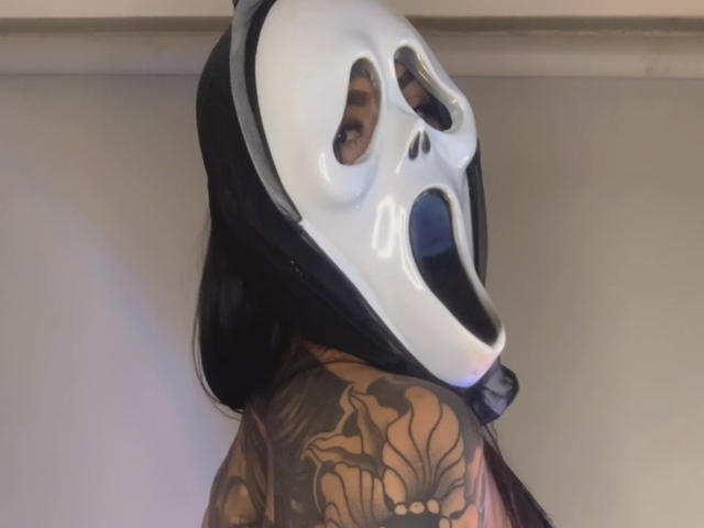Cylielee with a white, skull-faced mask, black head covering, and tattoos on her shoulder. Her mouth is open, showing her teeth. Cylielee has dark hair and a tattooed arm.