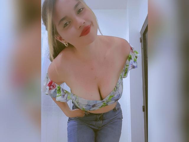 JulietaFoster69, leaning forward, wearing a floral off-shoulder top, exposing her big boobs, and blue jeans, with a slight smile, and red lipstick, in a bright room.