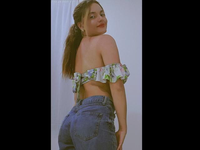 JulietaFoster69 with her ass in high-waisted blue jeans, wearing an off-shoulder floral top, looking over her shoulder with a slight smile, brown ponytail.
