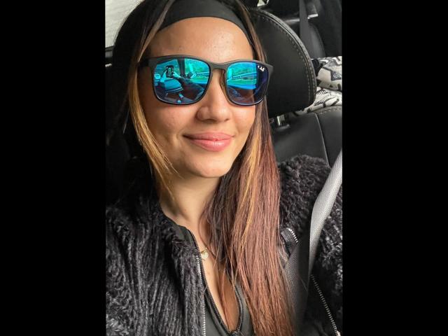 JulietaFoster69 with reflective blue sunglasses, black headband, and black textured jacket, sitting in a car, smiling with pink lips, brown and blonde hair.