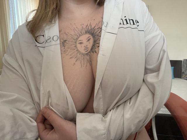 DiLaria with sun face tattoo between tits, wearing open white robe, holding it slightly, showing her chest and tattoo. Her tits are visible, she has light skin and brown hair.