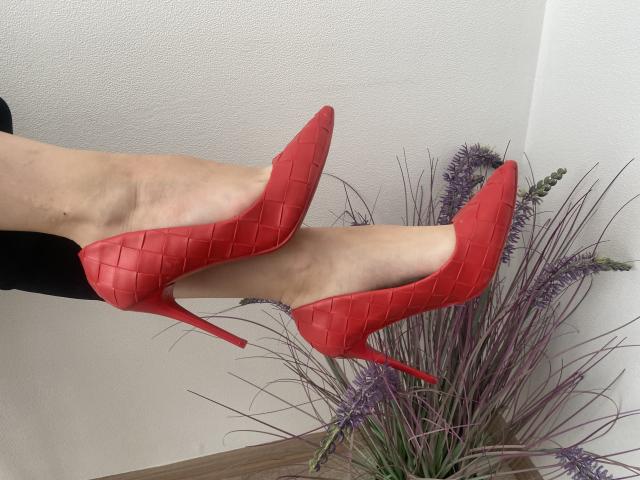 Vailetts Beine mit glänzenden, roten, krokodilmuster-verzierten Stiletto-Absätzen, die Füße im Knöchelbereich gekreuzt, mit einem schwarzen Outfit im Hintergrund. Eine lila Lavendelpflanze im unteren rechten Bereich..