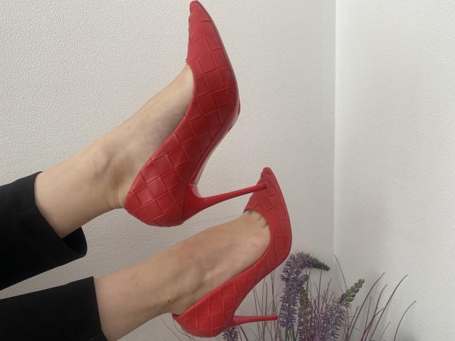 Vailett's legs in shiny red, high-heel stilettos with a woven pattern, showing off her bare feet and black pants. A purple plant is in the bottom right.