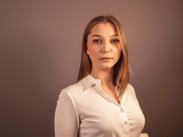 LovelyxEmilly with long brown hair, wearing a white button-up shirt, showing her tits, standing with a serious expression, against a plain brown background.