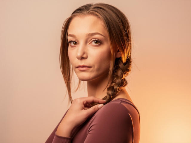 LovelyxEmilly with a side braid, wearing a purple top, looking over her shoulder, hand near her chin, soft orange lighting, smooth skin, subtle makeup, calm expression.