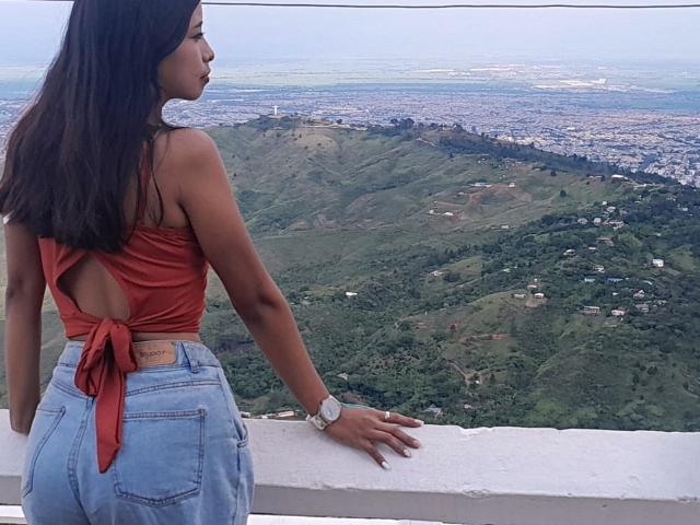 CaterinaRomano with long black hair, orange tied-up crop top showing ass, blue high-waisted jeans, standing against white railing, looking at green hill landscape, wearing white watch.