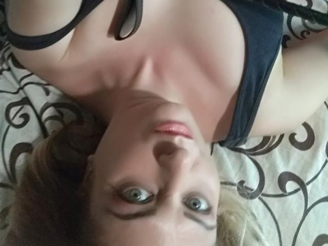 MonicaMiss lying on her back with blue eyes, black bra, and blonde hair, her face up, looking at the camera, her tits partially visible, on a patterned bed.