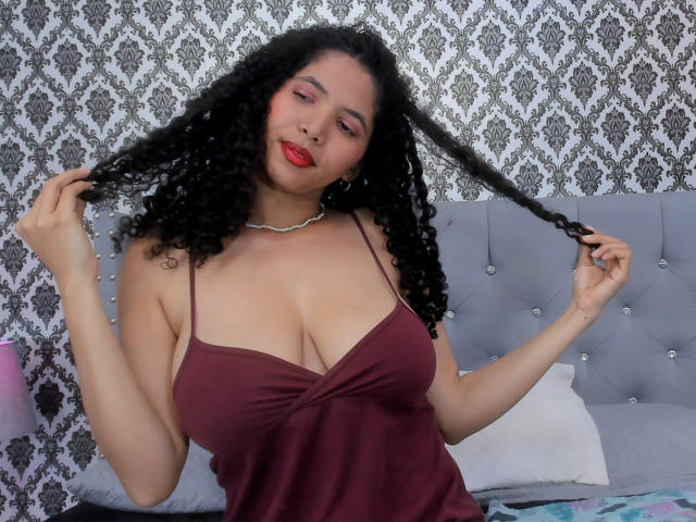 ZoeJonees with curly black hair, holding it, wearing a deep burgundy dress, showing her big boobs, red lipstick, and a silver necklace, sitting on a bed.