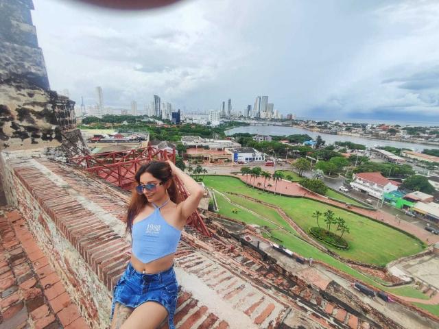 SaarahJones with tanned skin, brown hair, wearing blue crop top, blue denim shorts, and sunglasses, sitting on a brick ledge, city skyline and river in the background.