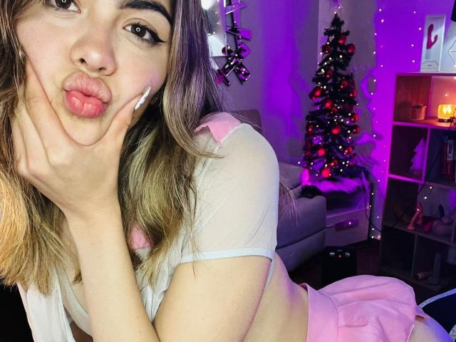 NathalySky with pouty lips, hands on cheeks, wearing a pink and white top, pink skirt, and long brown hair, in a purple-lit room with a decorated Christmas tree.