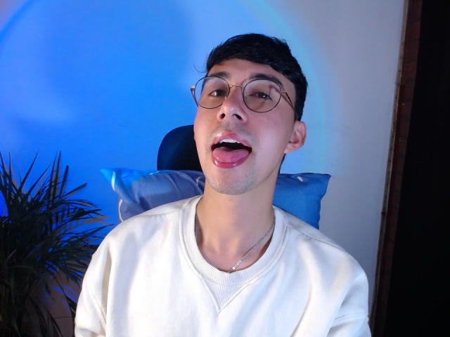 OliverHot69, with glasses and short black hair, tongue out, wearing a white V-neck, sitting against a blue-lit background, looking playful. Potted plant on left.