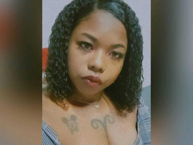 Baianna with curly black hair, brown skin, and tattoos above her tits, looking serious in a selfie. She's wearing a necklace and a checkered top.