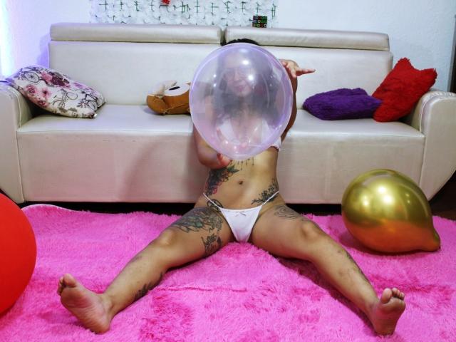 TinaFiery est assise sur un tapis rose, les jambes écartées, en train de souffler une bulle, elle porte des sous-vêtements blancs et a des tatouages sur les cuisses. Un ballon doré est à sa droite, et un canapé blanc se trouve derrière elle..