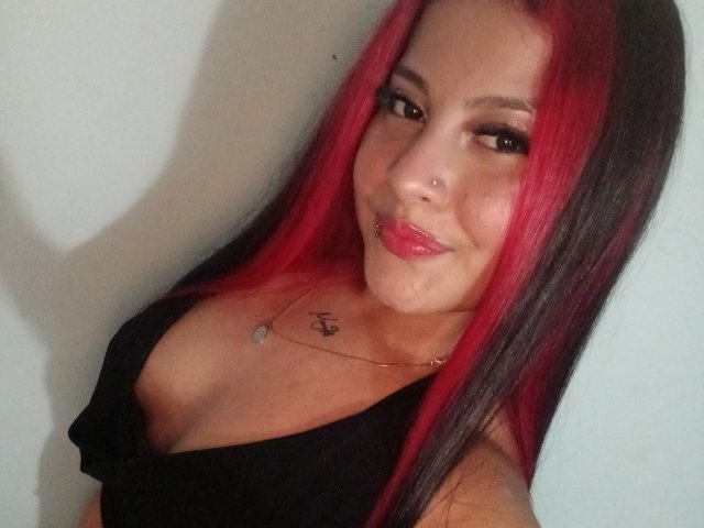 AshleyFostter69 with red and black hair, black top, nose ring, lip ring, tattoo on chest, smiling, medium tits, light skin, brown eyes, subtle makeup, against a white wall.