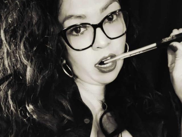 KarenCurlyHair with thick glasses, dark wavy hair, and hoop earrings, applying lipstick with a brush, wearing a dark button-up shirt, her lips slightly parted.