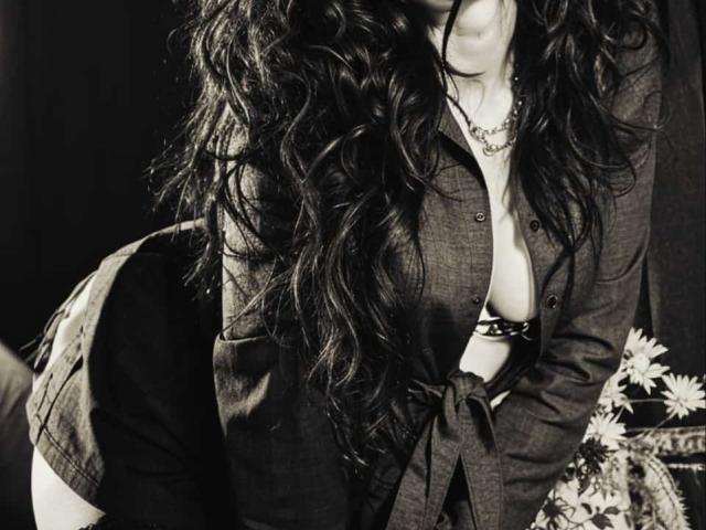 KarenCurlyHair with long, wavy dark hair, wearing an open button-down shirt revealing her big, bare boobs, black bra, and tied shirt, leaning forward with a flower bouquet.