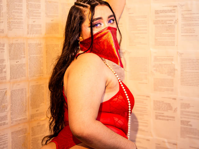 AishaGhulam with long wet black hair, red sheer lingerie, pearl necklace, and red velvet mask, showing her tits, standing against a wall, looking back with purple eyes.