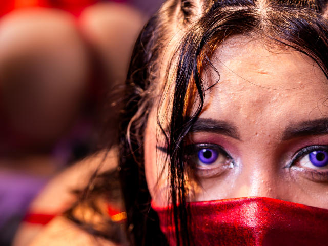AishaGhulam's face with wet black hair, purple eyes, and red mask, showing a slight sweat on her forehead. Her ass is blurred in the background.