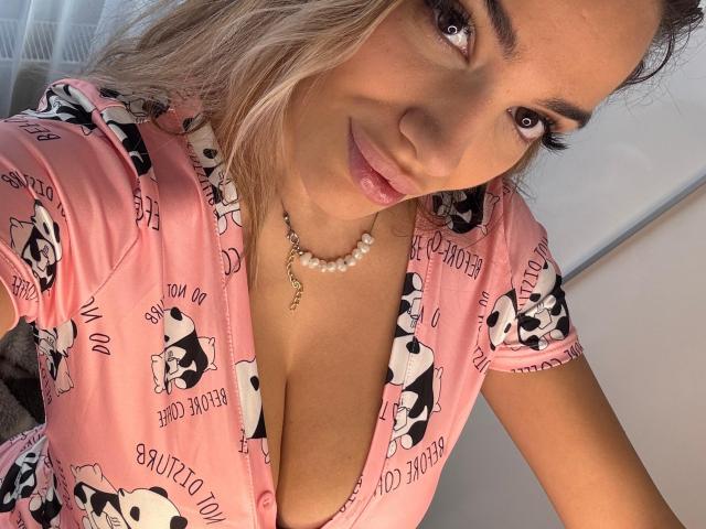 MiaNoirX has light brown skin, blonde wavy hair, and full pink lips. She's wearing a pink, dog-patterned dress with a deep V, showing her boobs. She has a pearl necklace.