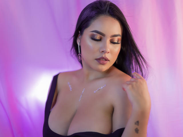 PaolaYKatty with dark hair, gold eyeshadow, and glitter on her tits, looking down with a serious expression, black strap dress, purple and pink background.