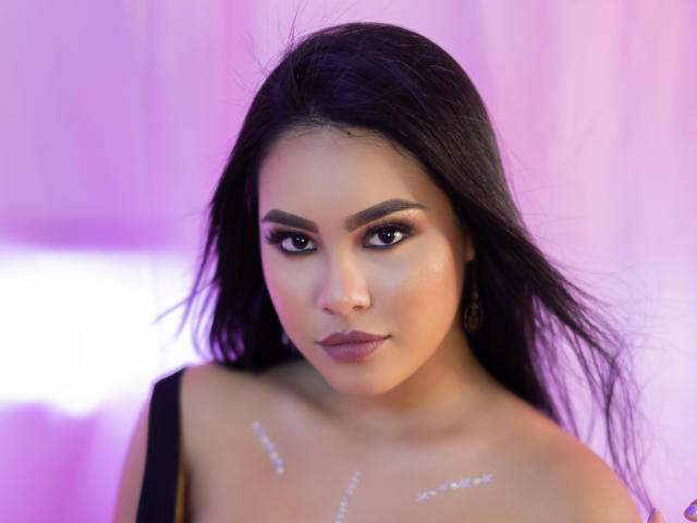 PaolaYKatty has long black hair, dark eyebrows, and subtle makeup. She's looking at the camera with a slight smile, showing off her small tits with glitter on her chest.